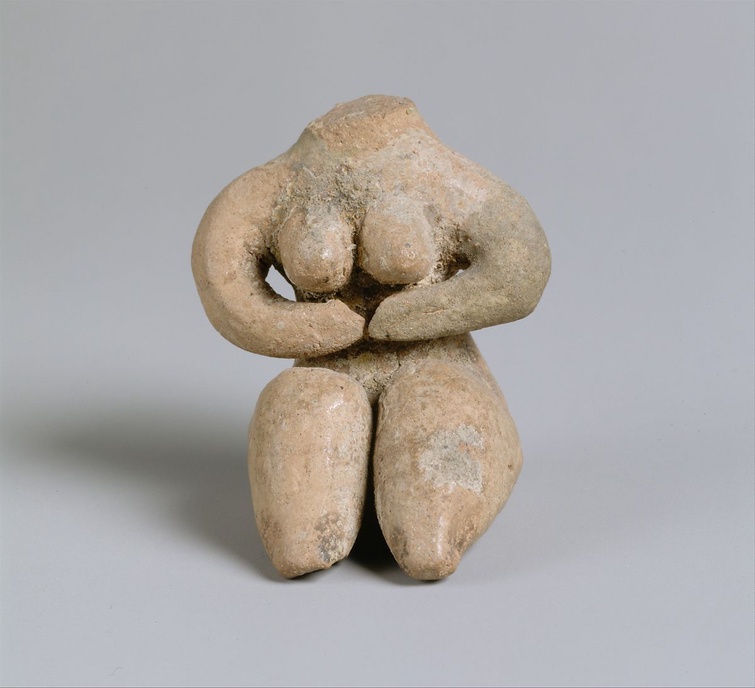 A small, ceramic, Halaf figure with traces of paint. The figure is headless but has prominent breast