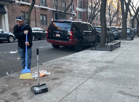 Person in black jacket and jeans with cigarette in mouth, sweeping trash with a blue broom on the si