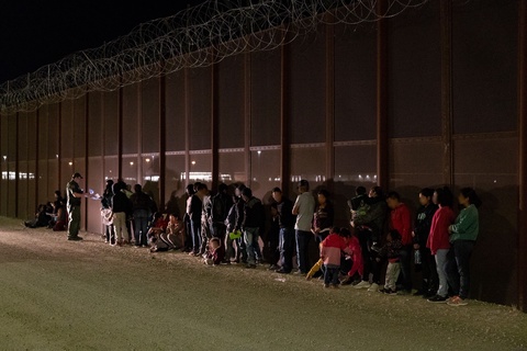 A US Customs and Border Protection agent is examining papers in front of a line of men, women and ch