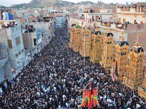 A street densely packed with people, most wearing black or dark colors. A row of tall, ornate gold p