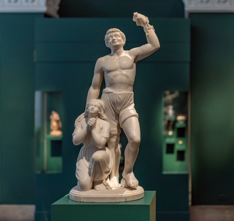 A photograph of a white marble sculpture in a green museum gallery. The sculpture consists of a stan