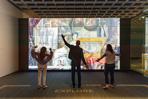 Three museum visitors are facing a large-scale illumination with their hands in various gestures bec