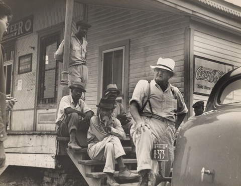 A black and white photograph features a white man dressed in a pale hat, shirt with rolled up sleeve