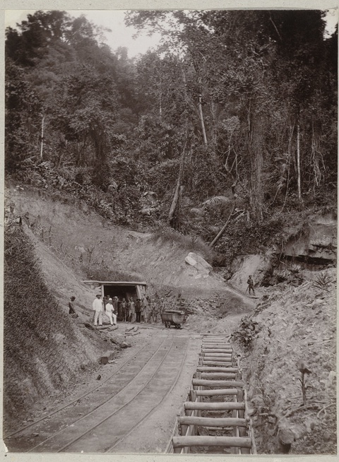 A pair of overseers, wearing clean white clothes, pose in front of a tunnel with a group of dark-ski