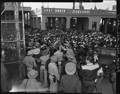 Huge crowd of men wearing hats fill an outdoor area flanked by a chainlink fence on the left and a v