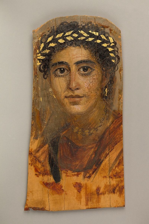 A portrait of a woman with dark curly hair and large round eyes. She adorns a gold wreath on her hea